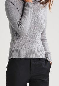 Ljust grå kabelstickad tröja med hög hals och ribbade ärmslut, kombinerad med svarta byxor. Stickad textur ger värme och detalj.