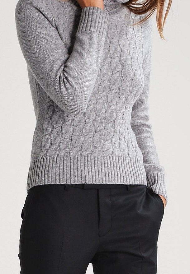 Ljust grå kabelstickad tröja med hög hals och ribbade ärmslut, kombinerad med svarta byxor. Stickad textur ger värme och detalj.