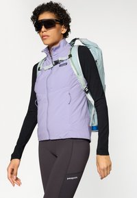 Vesta căptușită violet Patagonia, cu buzunare frontale, asortată cu un top negru cu mânecă lungă și colanți închiși la culoare, purtând un rucsac bleu deschis.