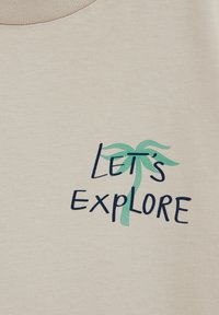 Béžová látka s tmavě modrým nápisem „Pojďme objevovat“ a zelenou stylizovanou palmou začleněnou do písmen.