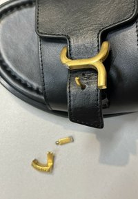 Gros plan d’une sandale en cuir noir avec une boucle métallique dorée cassée et deux pièces détachées de boucle dorée sur une surface blanche.