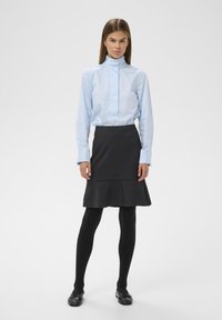 Chemise col bleu clair avec détails de boutons, jupe grise évasée, collants noirs et chaussures plates noires. Texture lisse, silhouette ajustée.