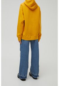 Personne portant un sweat à capuche jaune et un jean bleu à jambes larges avec des ourlets effilochés, debout de dos sur un fond clair uni.
