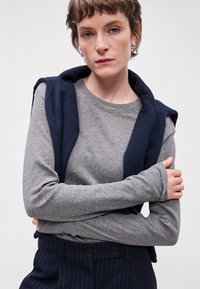 Camisola cinza de mangas longas com um decote redondo, sobreposta a uma peça externa sem mangas de cor azul marinho. A pessoa tem cabelo curto e usa brincos de argola.