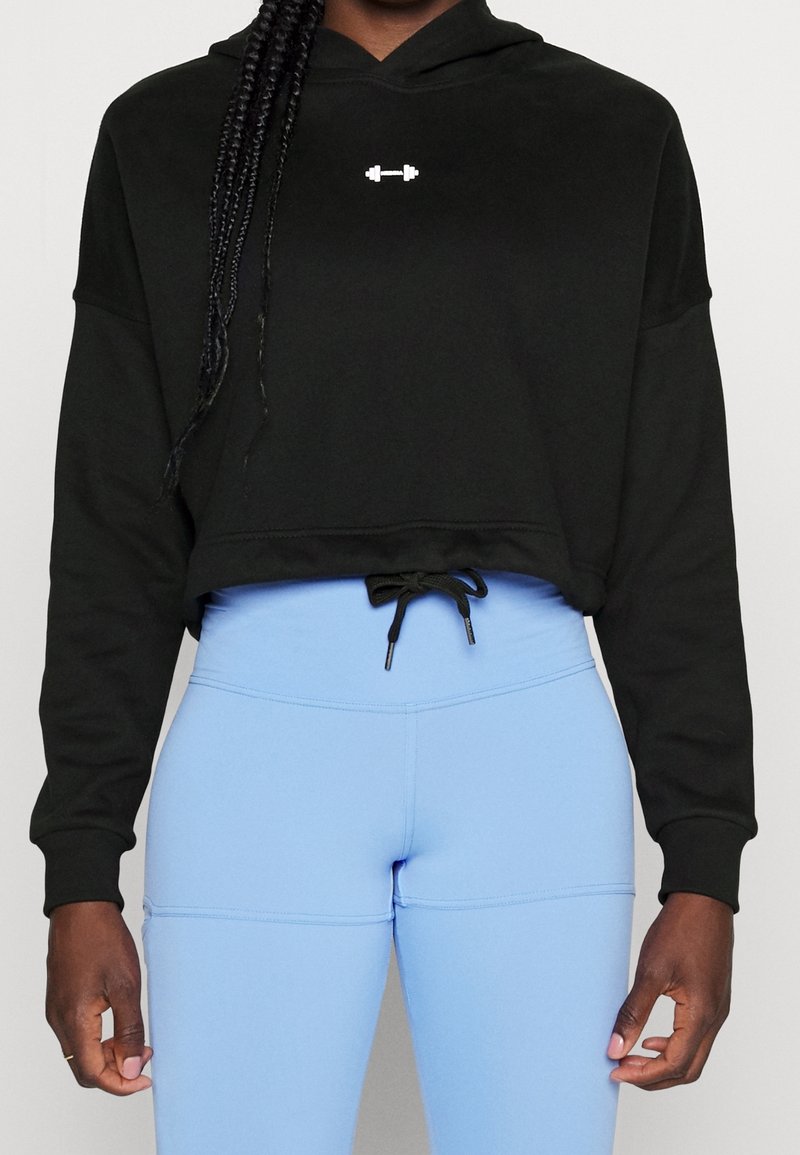 Personne portant un sweat à capuche court noir avec un petit logo barre blanche et des leggings bleu clair taille haute avec un cordon noir.