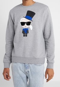 Persona che indossa un felpa grigia con una figura dei cartoni animati in occhiali da sole, un cilindro e un papillon blu stampati sul davanti.