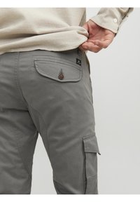 Personne ajustant une chemise beige rentrée dans un pantalon cargo gris avec une poche à rabat boutonnée et une étiquette noire sur la poche arrière.
