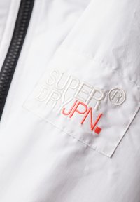 Tissu de veste blanche avec de subtiles rayures verticales. Présente un logo brodé en gris et orange : "SUPER DRYJPN." Fermeture à glissière visible.