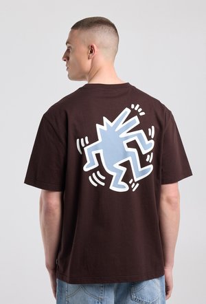 Man met een kort kapsel die een donkerbruin T-shirt draagt met een grote blauwe en witte abstracte dansfiguur op de rug, staand opzij.