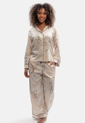 Woman with curly hair smiling, wearing beige satin pajamas with abstract line art patterns and black piping, standing with one hand in pocket.