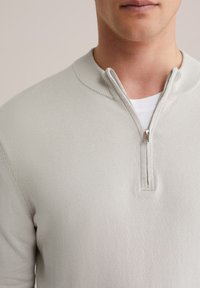 Pull en tricot gris clair avec un col à fermeture éclair, doté de détails côtelés et d'une texture lisse. Porté sur une couche blanche en dessous.