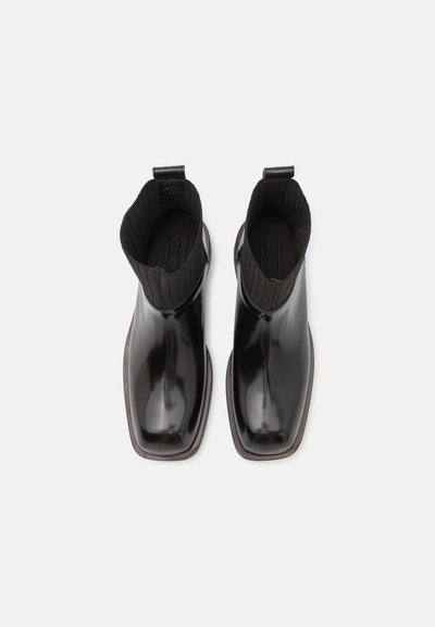 Botas de tornozelo pretas e brilhantes com um colar de malha canelada, bico quadrado e um salto discreto. Sem hardware ou detalhes visíveis. Textura suave.