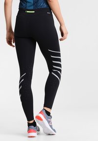 Leggings atletici neri con un design aderente, caratterizzati da strisce riflettenti sui lati e una cintura testurizzata. Adatti per l'abbigliamento sportivo.