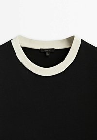 Chemise noire avec un col rond ivoire et une étiquette de vêtement visible à l'intérieur de l'encolure.