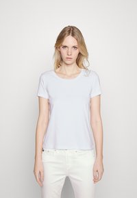 Camiseta de algodón blanca con cuello redondo y mangas cortas. Presenta un detalle de logo impreso en el hombro izquierdo. Textura suave.