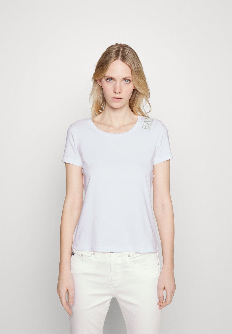 Camiseta de algodón blanca con cuello redondo y mangas cortas. Presenta un detalle de logo impreso en el hombro izquierdo. Textura suave.