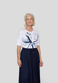 Weißes T-Shirt mit abstraktem blauen Grafikdesign, Rundhalsausschnitt und kurzen Ärmeln, kombiniert mit einem marineblauen Faltenrock mit elastischem Bund.