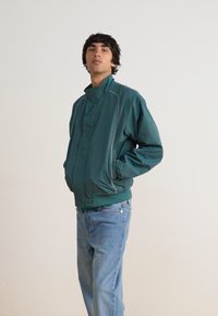 Turkizna lahka jakna z visokim ovratnikom, sprednjim zadrgo in belimi detajli. Nosi se nad svetlo modrimi jeans hlačami. Enostavna ozadje.