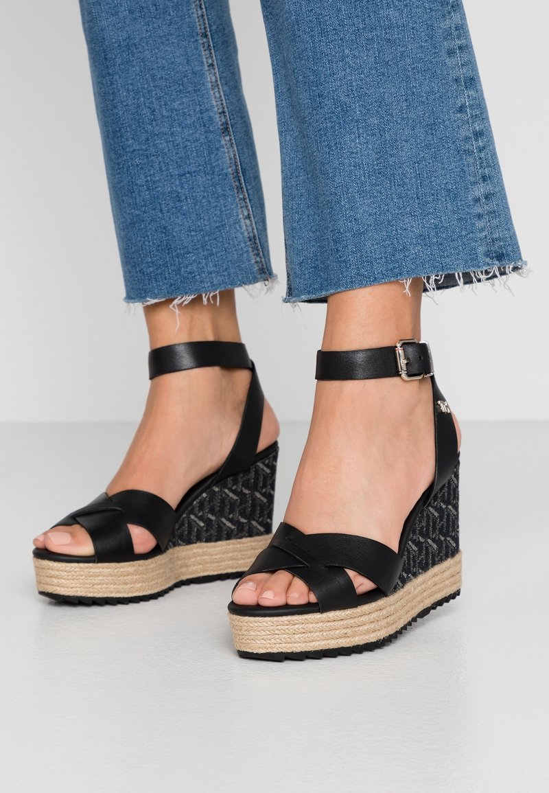 Pieds portant des sandales compensées en cuir noir avec bride cheville, semelles plateformes en jute tressé et jeans denim bleu coupés courts avec ourlets effilochés.