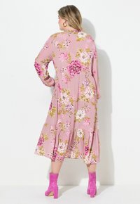 Rotes Kleid mit langen Ärmeln, das mit großen weißen und lila Blumen verziert ist, mit einem gestuften Saum, getragen mit rosa Absatzstiefeln.