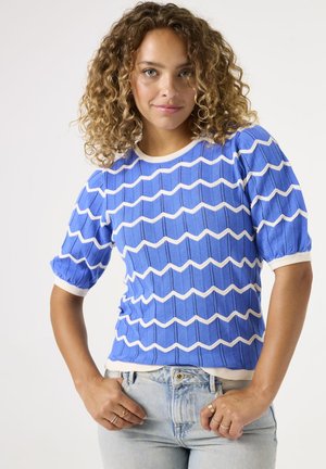 Blauw kortemouwen trui met een golvend wit patroon, ribbelstructuur en een witte halslijn. Gecombineerd met lichtblauwe spijkerbroek.