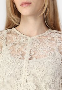 Haut en dentelle beige avec un motif floral et un design texturé. Il présente un col rond et une fermeture à boutons à l'avant, avec des sections transparentes.