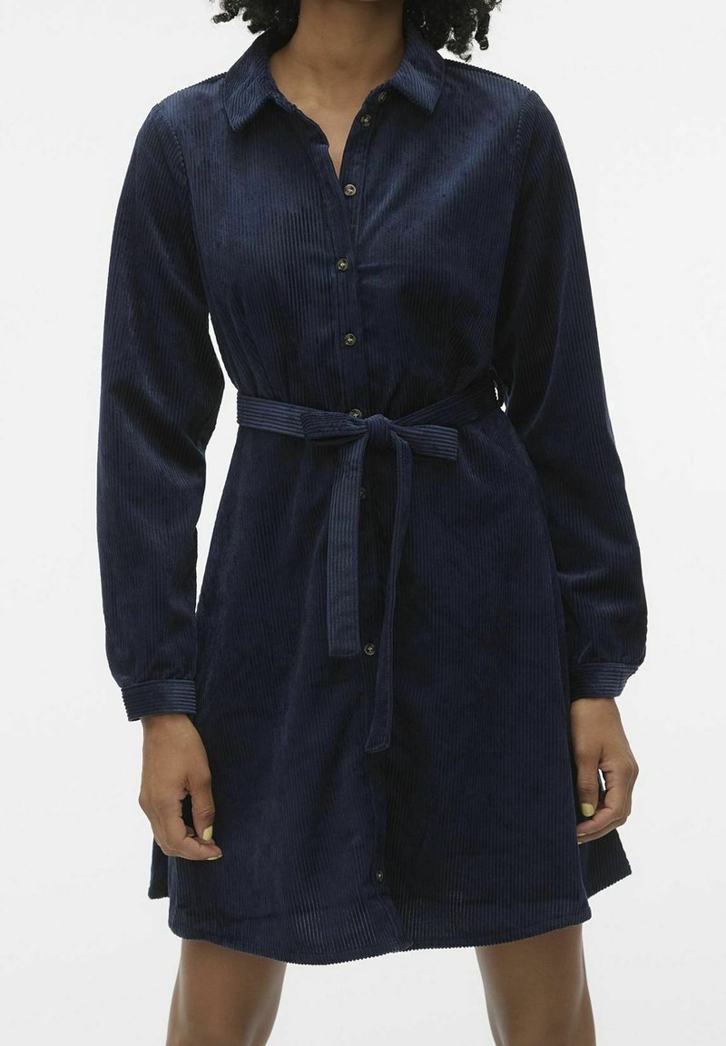 Femme portant une robe chemise en velours côtelé bleu marine avec manches longues, boutonnée à l'avant, col et ceinture nouée à la taille.