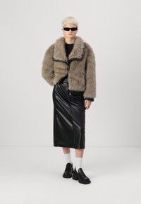Donkerbruin faux fur cropped jack met brede kraag, gecombineerd met een zwarte leren midi-rok. Het model draagt zwarte zonnebril en stevige zwarte schoenen.