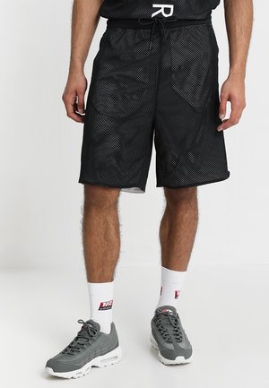 Mężczyzna w czarnych siateczkowych szortach, białych skarpetkach z czerwonym logo i szarych sportowych butach, stojący na białym tle.