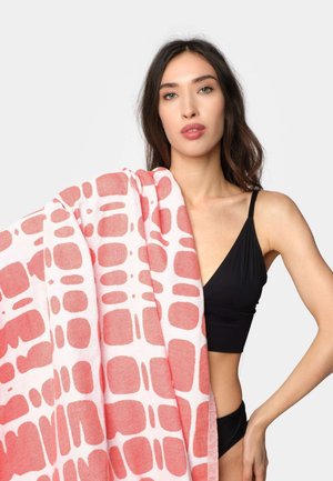 Donna in bikini nero che tiene una grande asciugamano rosa e bianca a motivo sulla spalla contro uno sfondo semplice.