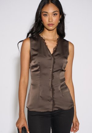 Woman wearing sleeveless brown satin blouse with lace trim and black pants, holding a black clutch in her left hand.