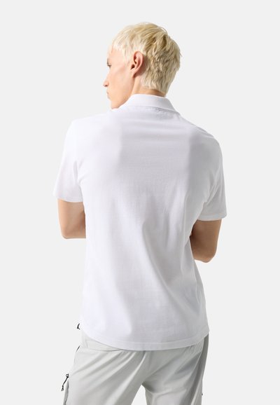 Young man with short blond hair wearing a white polo shirt and light gray pants, facing away against a plain light background.