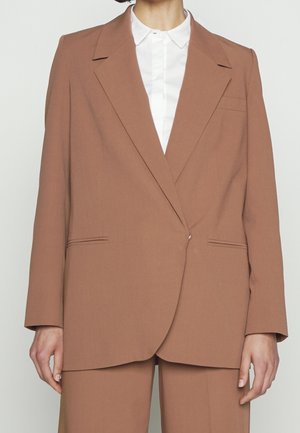 Femme portant un blazer marron ajusté avec un pantalon assorti et une chemise blanche à col, debout devant un fond clair uni.