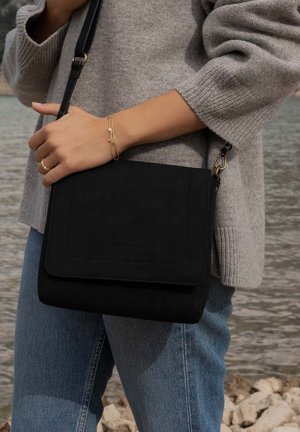 Zwarte suede schoudertas met een klepsluiting, gekenmerkt door een gladde textuur en een verstelbare riem, gecombineerd met een grijze gebreide trui en blauwe spijkerbroek.