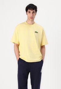 Yellow cotton t-shirt with a crew neck, short sleeves, and an embroidered green crocodile logo on the left chest. Paired with navy pants.