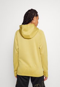 Frau mit geflochtenem Haar, die einen gelben Kapuzenpullover und schwarze Shorts mit gelber "WK"-Beschriftung trägt, von hinten vor einem einfarbigen Hintergrund.