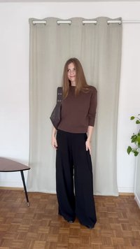 Un pull marron côtelé associé à un pantalon large noir, un sac à main sombre, se tenant sur un sol en bois devant un rideau clair.