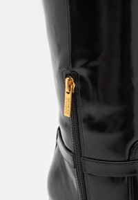 Stivale in pelle patent nera con zip laterale e inserto dorato, caratterizzato da cuciture a contrasto. Texture liscia e design strutturato.