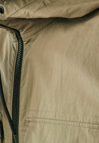 Veste tan en tissu lisse et léger, avec fermeture éclair à l'avant et une poche sur le côté gauche avec des coutures contrastantes.