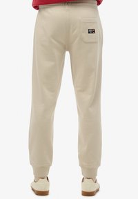 Beige mjukisbyxor med elastisk midja och muddar. Har en ensam bakficka med en logotagg. Slät textur, avslappnad passform.