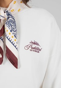 Donna che indossa un felpa bianca con ricamo "Proibito 10119 Berlino" e una sciarpa di seta stampata annodata attorno al collo.