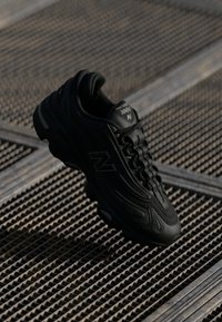 Chaussure de sport noire avec des surfaces textureées, des courbes prononcées et la marque New Balance. Elle dispose d'une semelle rembourrée et d'un système de laçage.