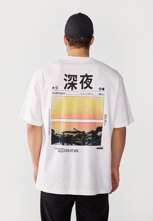 Homme portant un T-shirt blanc avec des caractères japonais noirs et un motif de paysage de coucher de soleil dans le dos, casquette noire et pantalon foncé.