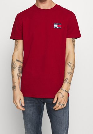Mężczyzna noszący czerwoną koszulkę Tommy Jeans i niebieskie dżinsy, pokazujący liczne tatuaże na obu ramionach i dłoniach.