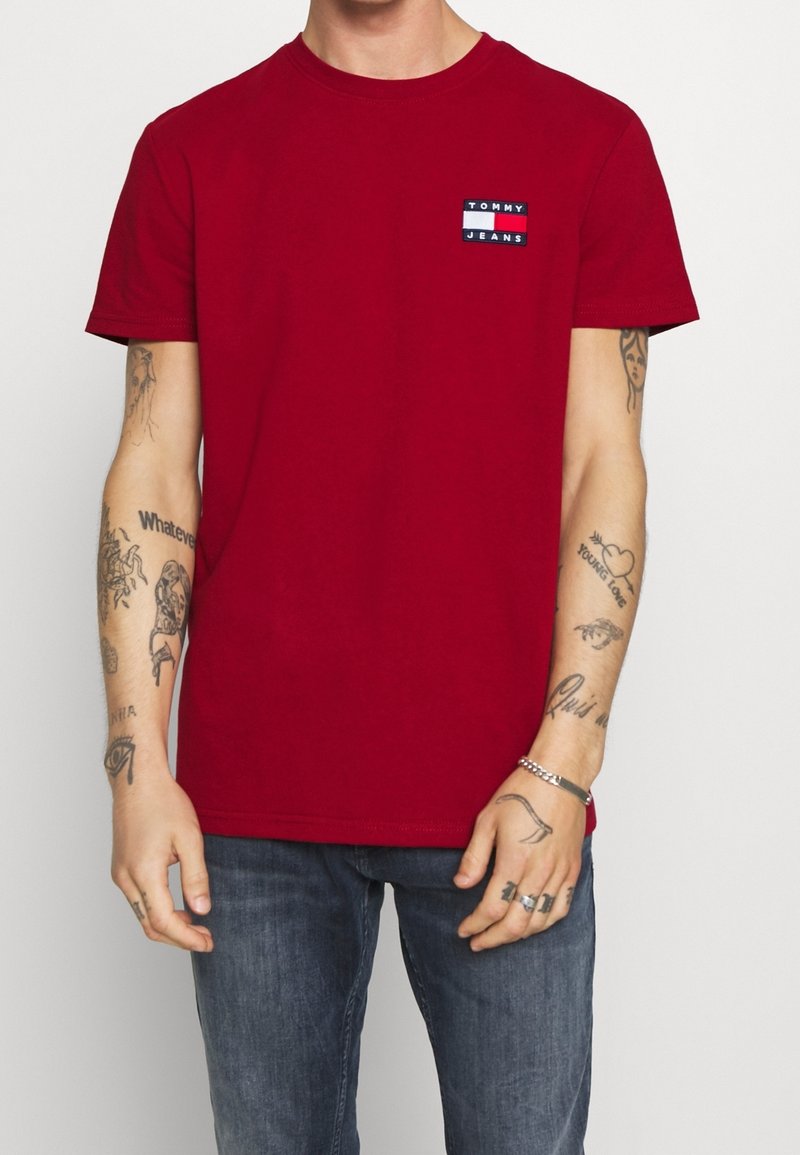 Homme portant un t-shirt rouge Tommy Jeans et un jean bleu, montrant plusieurs tatouages sur les deux bras et les mains.