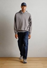 Grå hettegenser, blå jeans og grå joggesko; person med en mørk cap, i avslappet passform og myk tekstur. Flat bakgrunnsoverflate.