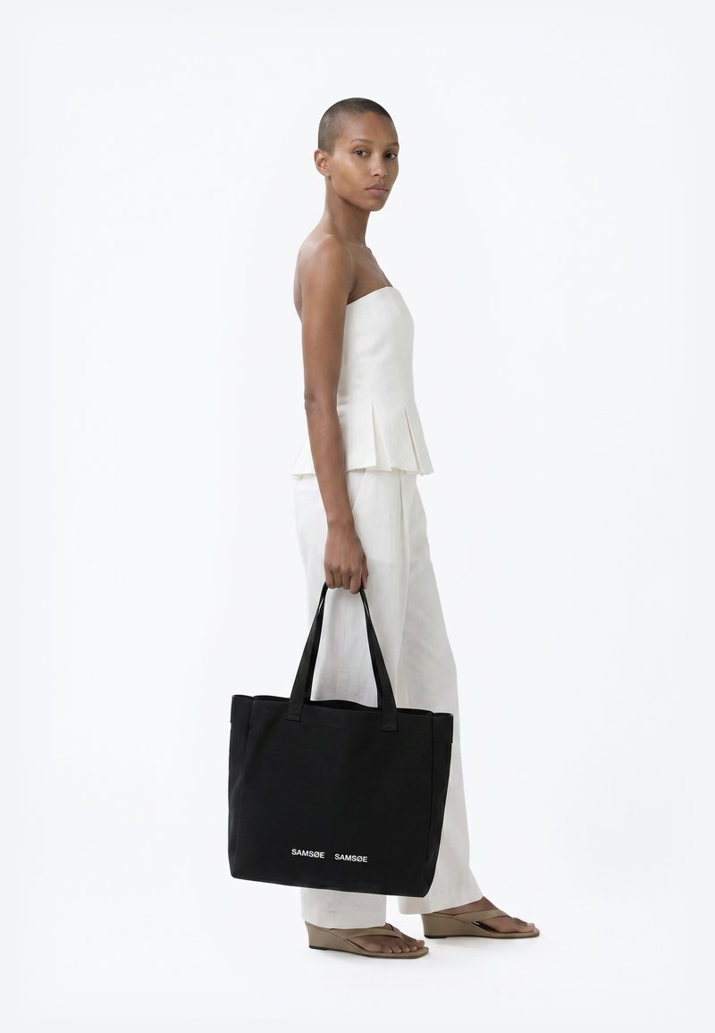 Femme en haut blanc sans manches et pantalon tenant un sac cabas noir avec le texte "SAMSØE SAMSØE", debout sur fond blanc.