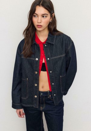 Ung kvinde iført mørk denimjakke og jeans over en rød cropped skjorte, stående mod en ensfarvet lys baggrund.