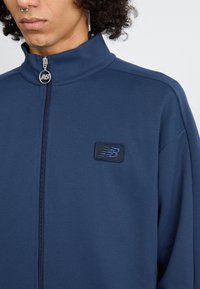 Navy blue zip-up jacket made from textured fabric, featuring a high collar and a small black and blue New Balance logo on the chest.