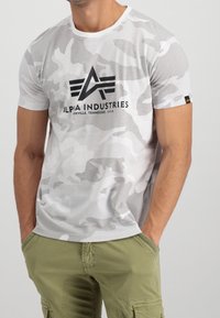 Camiseta de manga corta blanca con patrón de camuflaje gris; presenta un logo negro "ALPHA INDUSTRIES" en el pecho; textura suave.